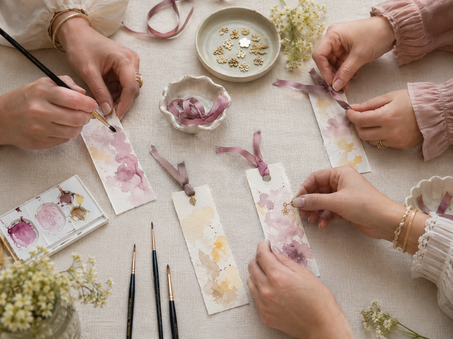 Women’s hands painting watercolor bookmarks and adding mauve ribbons and small gold charms at a craft table